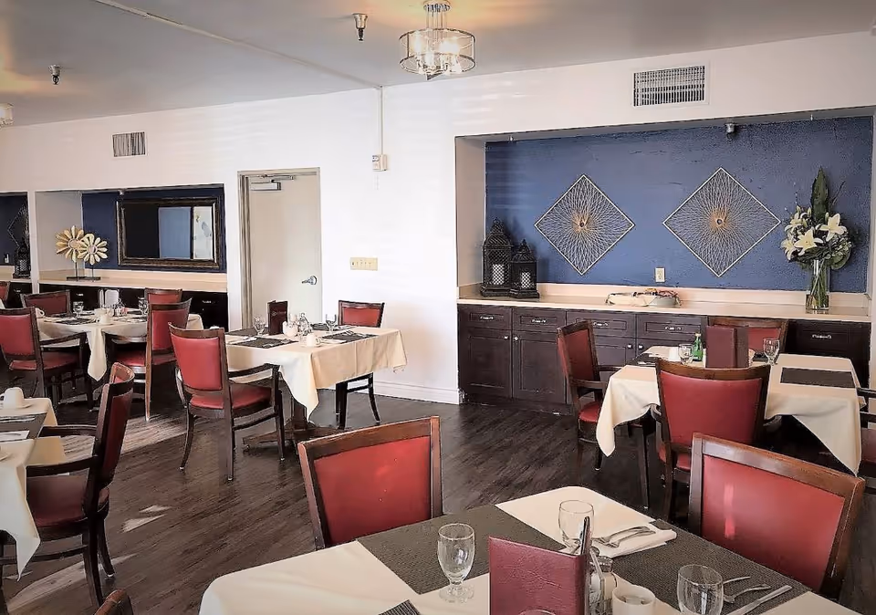 Dining room with tables set with white tablecloths and red chairs, dark wood cabinetry and decorative wall art.