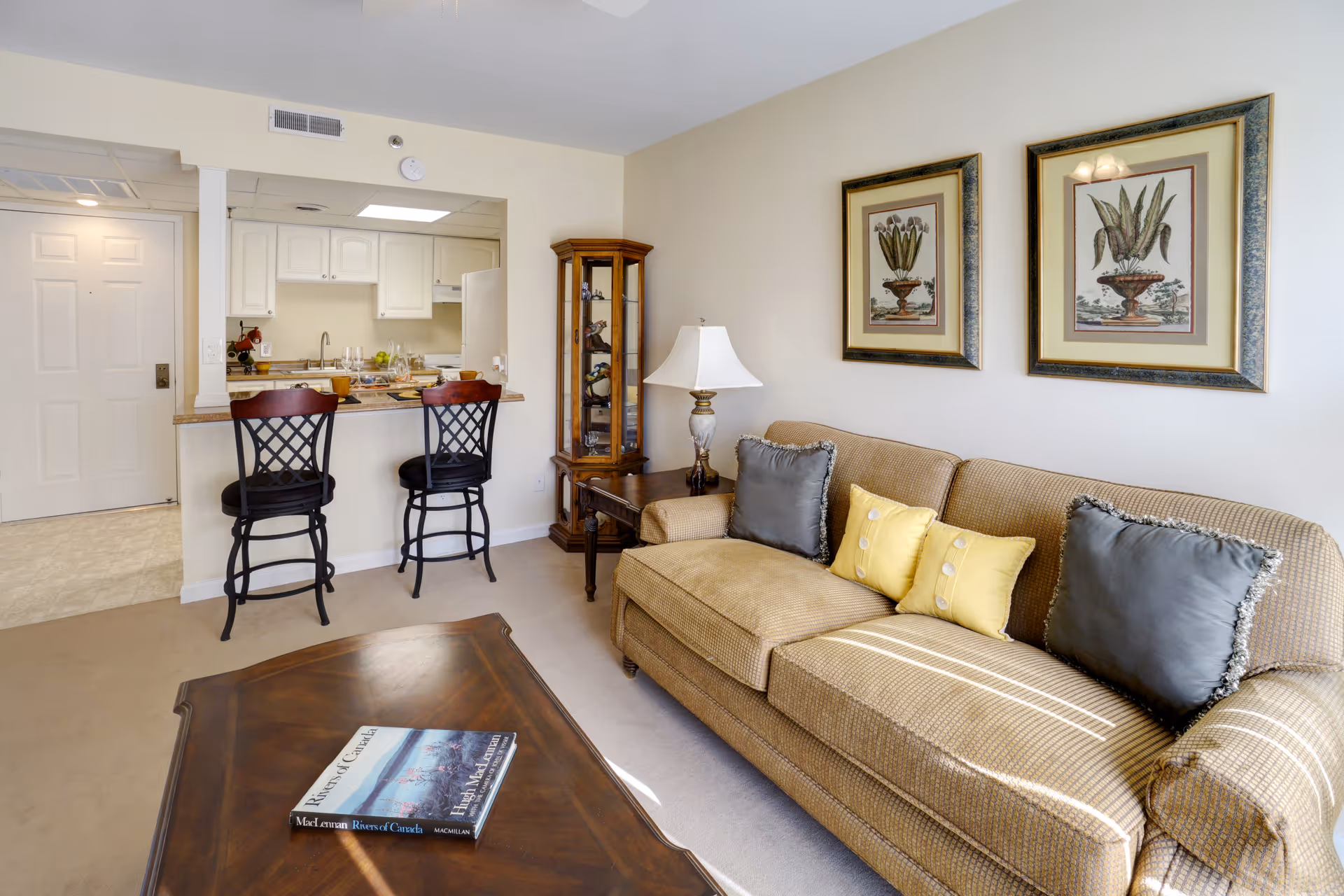 A cozy living room area with a beige couch adorned with two gray and two yellow pillows. Above the couch are two framed botanical prints. To the right of the couch is a wooden side table with a lamp and a tall glass display cabinet. In front of the couch is a wooden coffee table with a book on it. The living room opens to a kitchen area with a counter and two high chairs. The kitchen has white cabinets and appliances. A white door is visible in the background.