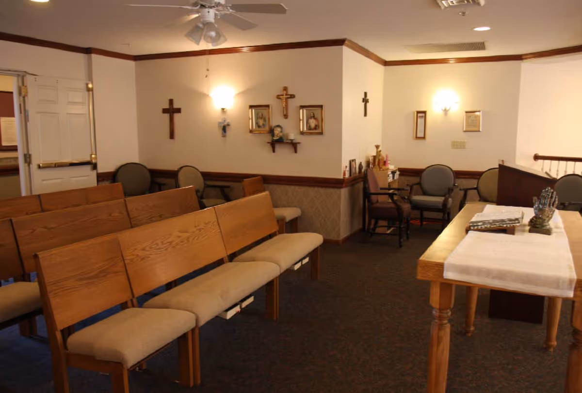A small chapel or prayer room with wooden pews and cushioned seats arranged facing a wooden altar table covered with a white cloth. The walls are decorated with several crosses and religious pictures. There are additional chairs along the walls and soft lighting from wall sconces and a ceiling fan light.