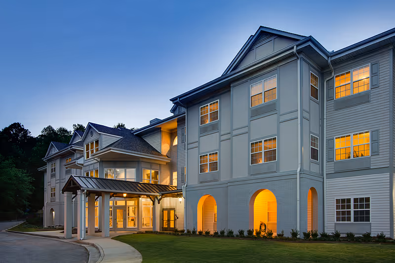 Exterior view of a multi-story senior living facility building at dusk with warm lights glowing from the windows. The building has a covered entrance with columns and a driveway leading up to it, surrounded by a well-maintained lawn and trees in the background.