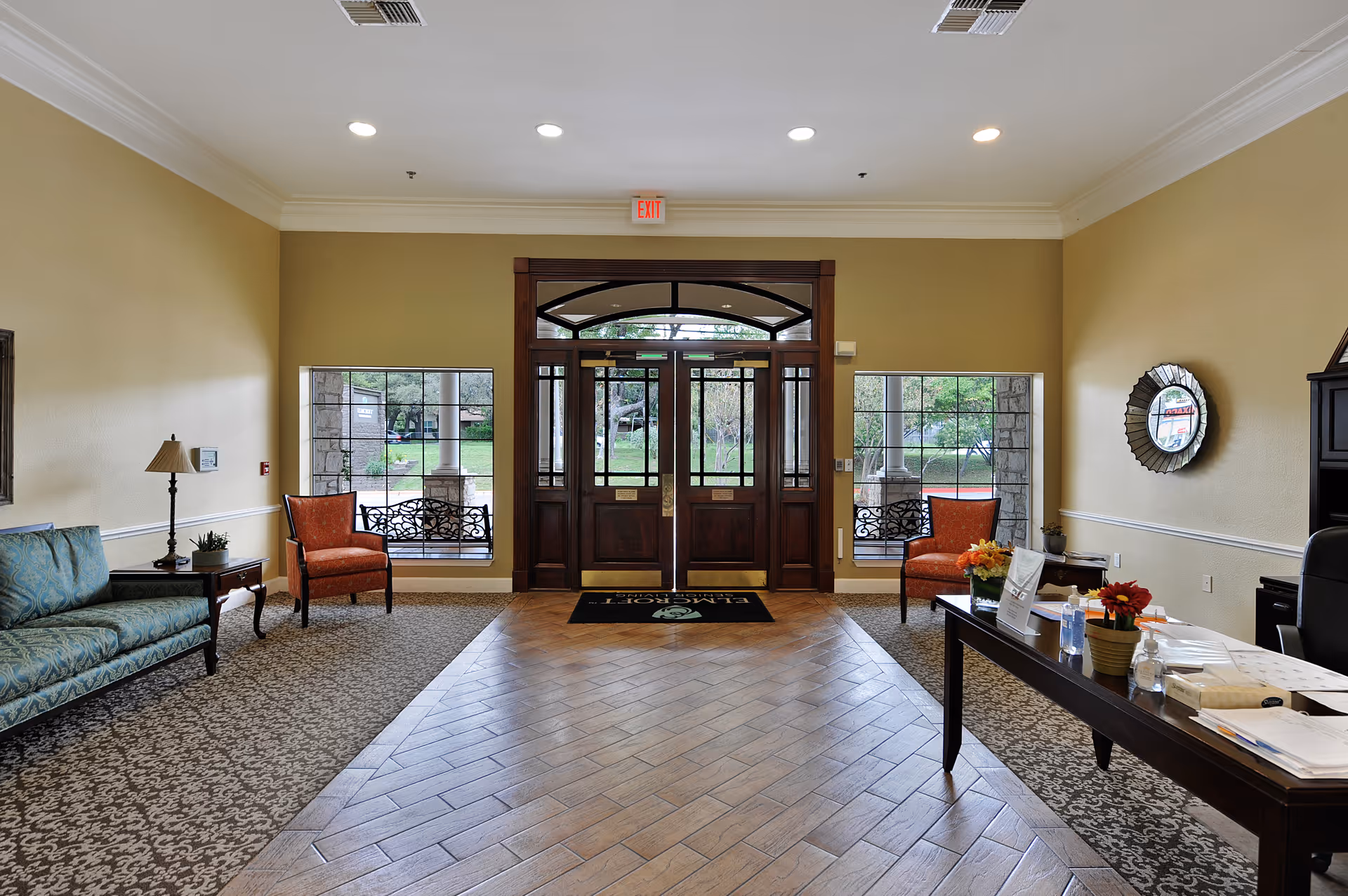 Bright entrance lobby with double wooden doors, seating areas on either side, and a reception table on the right.