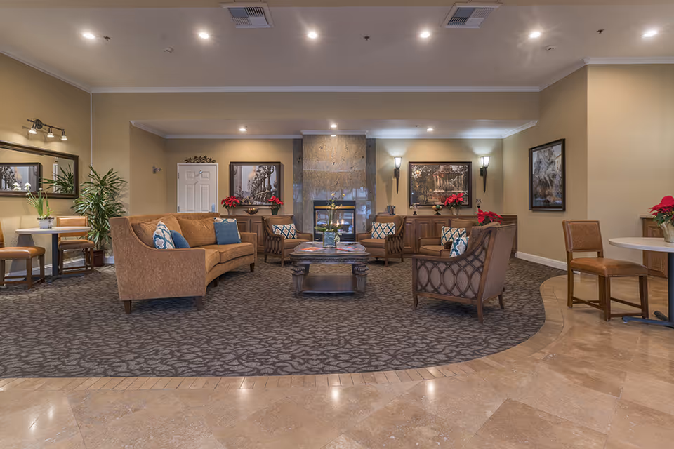 A cozy senior living facility common area with a central fireplace surrounded by brown cushioned chairs and sofas with blue and patterned pillows. The room features beige walls adorned with framed artwork, potted plants, and red poinsettia flowers. There are small tables and chairs on the sides, and the floor is a combination of patterned carpet and tile.