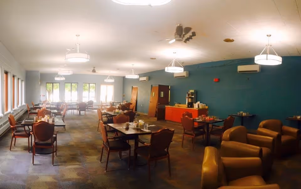 A spacious dining room with multiple tables and chairs arranged neatly. The room has large windows on one side allowing natural light to enter. There are several ceiling lights and ceiling fans. A counter with a coffee machine and supplies is against a teal-colored wall. Comfortable armchairs are placed near the corner of the room.