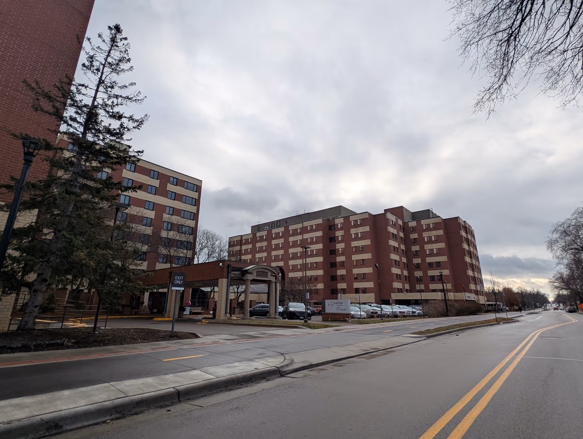 Exterior view of a large multi-story brick building with multiple windows, a covered entrance, and a parking lot with cars. There is a sign near the entrance that reads 'Walker Place'. The sky is cloudy and the street in front of the building is empty.