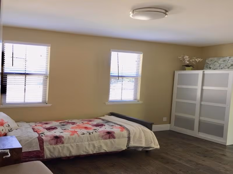 Well-lit bedroom with a single bed with floral bedding, two windows with blinds, and a white wardrobe against a beige wall.