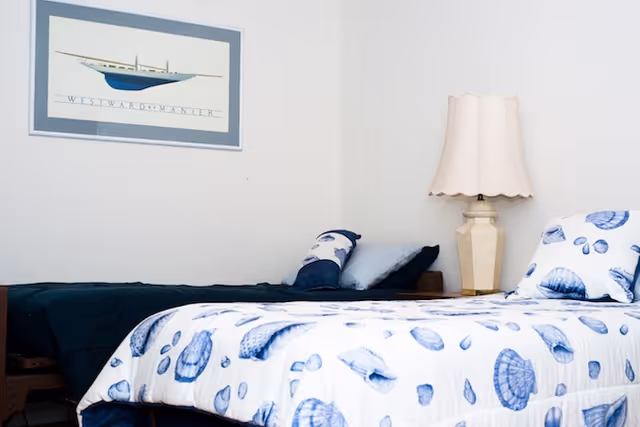 A bedroom with two single beds, one with a white bedspread featuring blue seashell patterns and the other with a dark blue bedspread. There are pillows on each bed, a beige table lamp on a small nightstand between the beds, and a framed picture of a boat on the wall above the beds.