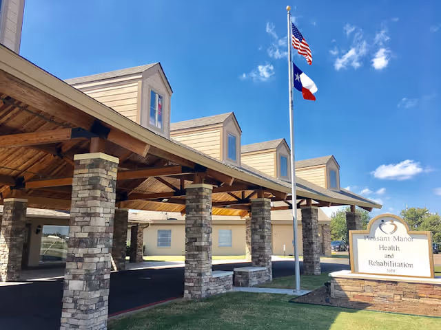 Exterior view of Pleasant Manor Health and Rehabilitation building with a covered entrance supported by stone pillars, an American flag and Texas state flag flying on a flagpole, and a clear blue sky with a few clouds.