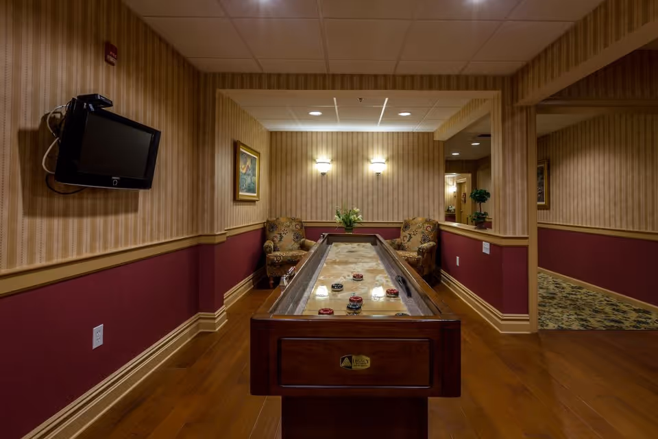 A cozy indoor recreational room with a shuffleboard table in the center, two patterned armchairs at the far end, a wall-mounted TV on the left, and warm lighting from wall sconces. The walls have striped wallpaper with a maroon lower half and wooden trim, and the floor is hardwood.