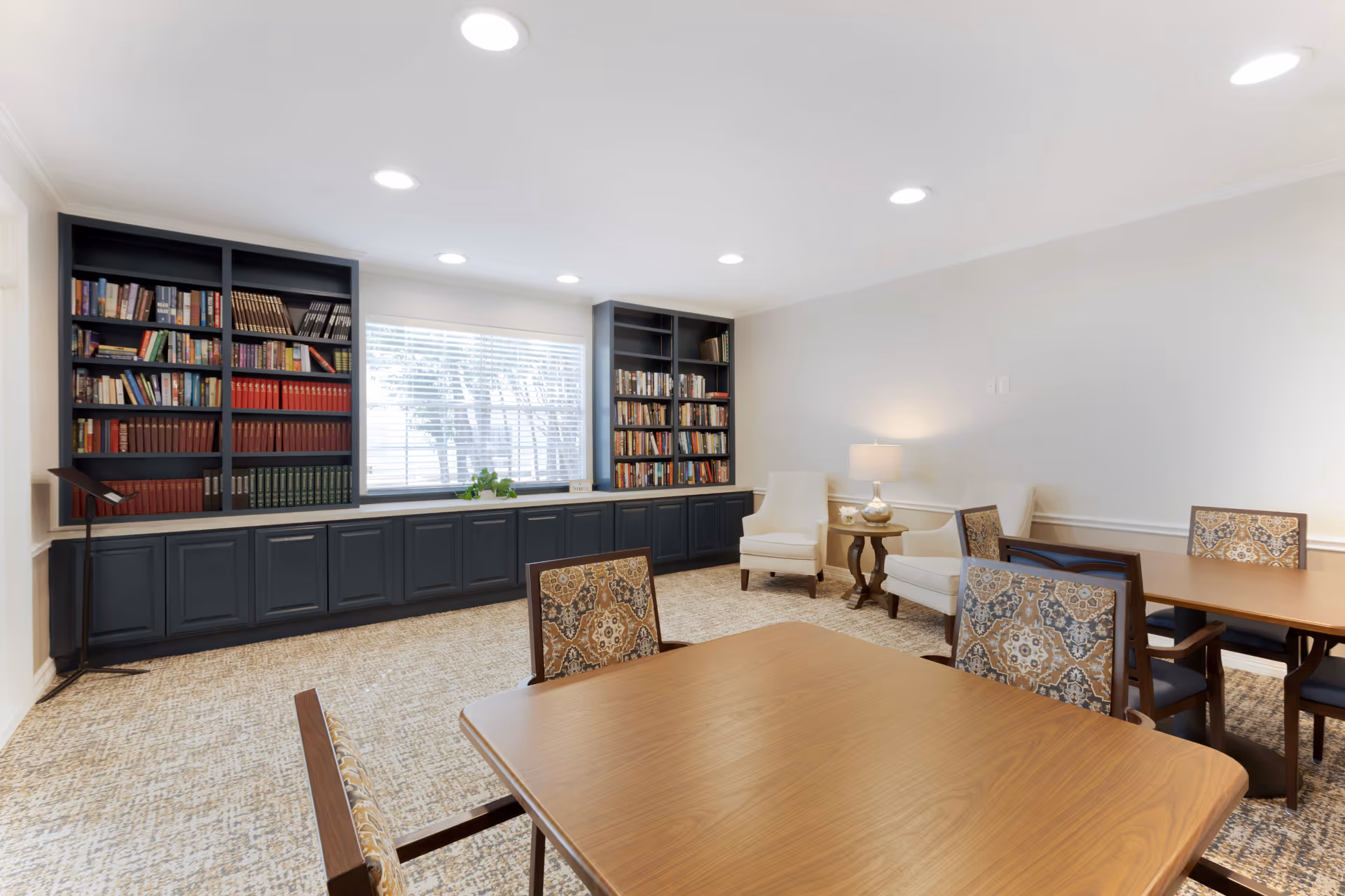 A bright and cozy senior living common room with wooden tables and patterned chairs. There are two beige armchairs with a small round table and lamp between them. The back wall features built-in dark blue bookshelves filled with books and a large window with blinds letting in natural light. The room has recessed ceiling lights and a beige patterned carpet.