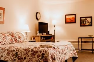 A cozy bedroom with a floral patterned bedspread on a bed, two folded towels placed on the bed, a small TV on a stand, a clock on the wall, two framed pictures, and a floor lamp providing warm lighting.