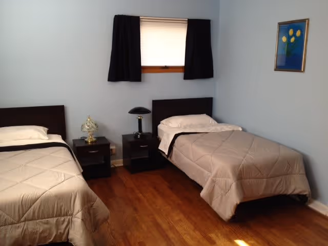 A simple bedroom with two single beds, each with beige comforters and white pillows. Between the beds are two dark wooden nightstands, one with a decorative lamp and the other with a black lamp. A small window with black curtains is centered on the wall behind the beds. A framed picture with yellow flowers hangs on the right wall. The floor is wooden.