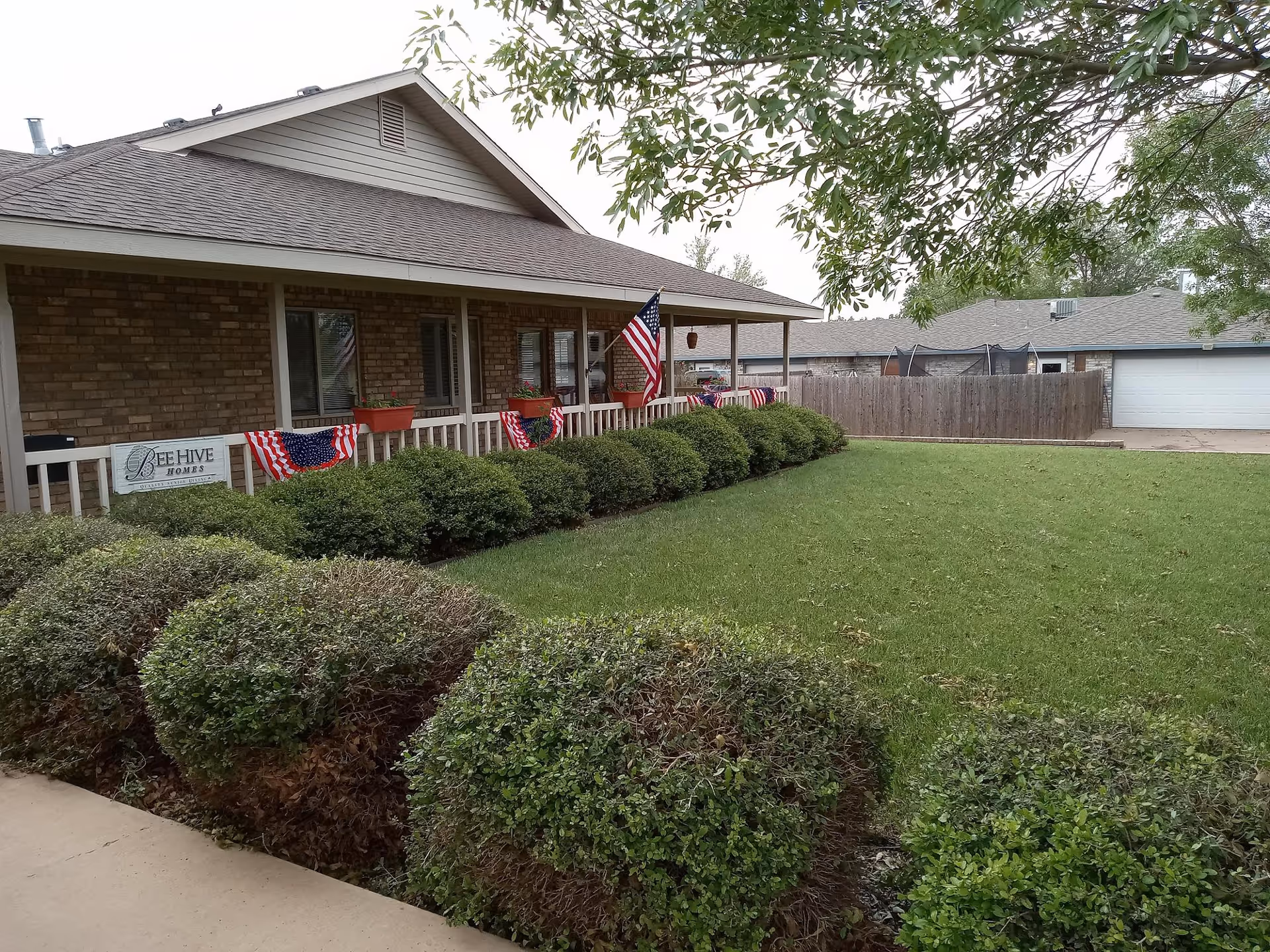 Front exterior of a BeeHive Homes building showing a covered porch decorated with American flags and a manicured lawn.