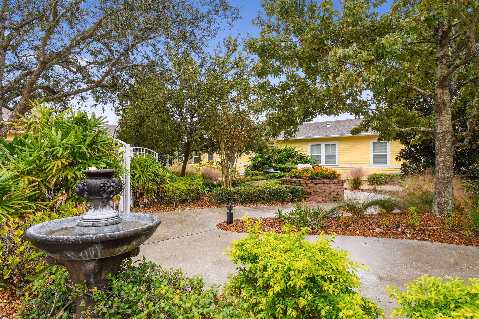 A landscaped outdoor garden area with a stone water fountain in the foreground, surrounded by green shrubs and trees. A curved concrete pathway leads through the garden, with a yellow building with white-trimmed windows visible in the background under a blue sky.