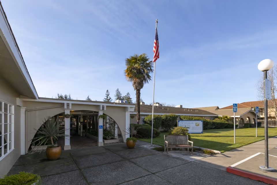 Exterior view of Broadway Villa Post Acute facility showing the entrance with an archway, potted plants, a bench, a flagpole with an American flag, and a lawn area with a sign. The sky is clear and blue.