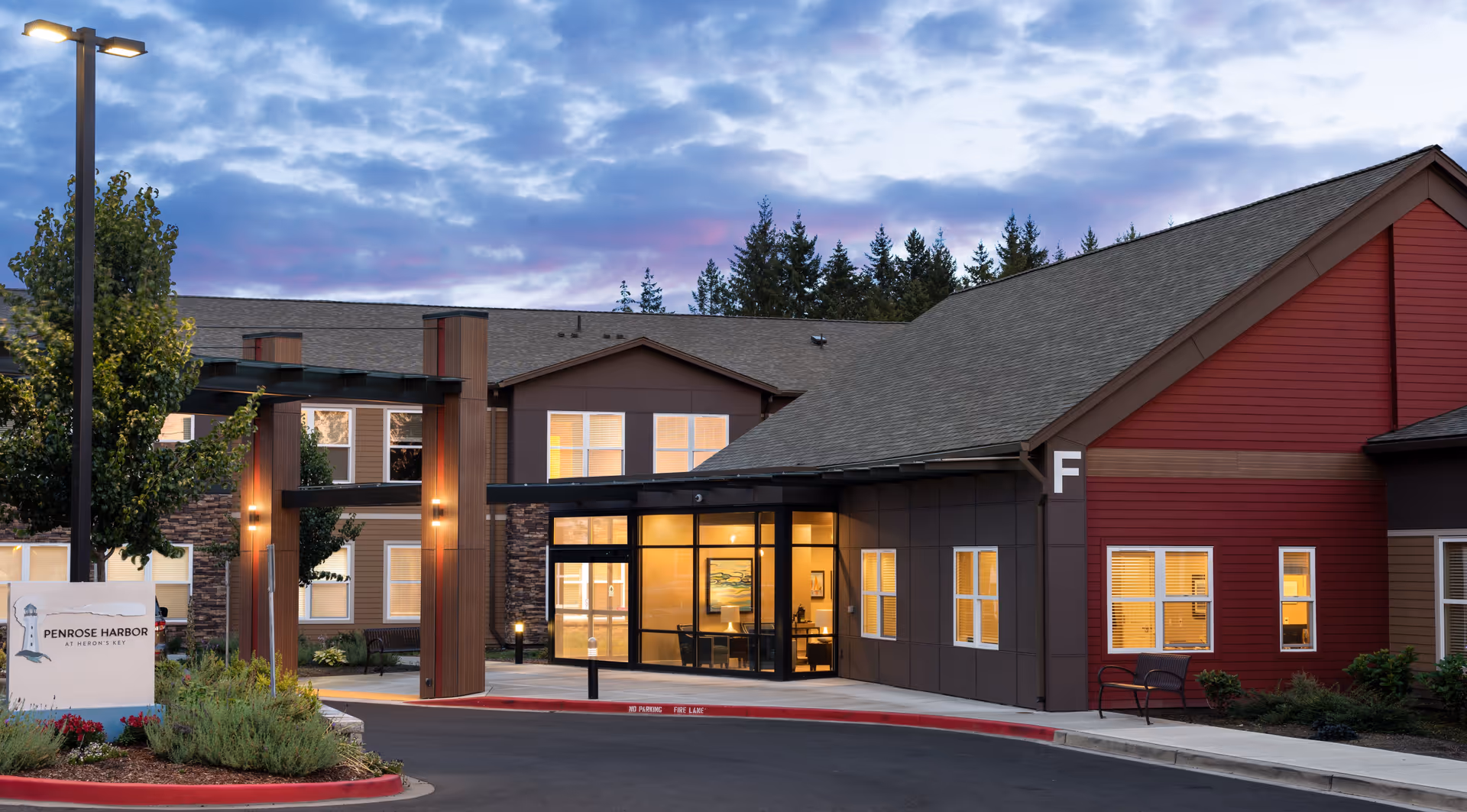 Exterior view of a senior living facility building at dusk with warm interior lights visible through large windows. The building has a combination of brown, red, and beige siding with a covered entrance supported by wooden pillars. A sign near the entrance reads 'Penrose Harbor at Heron's Key'. Trees and landscaping surround the area, and a streetlamp is lit near the entrance.