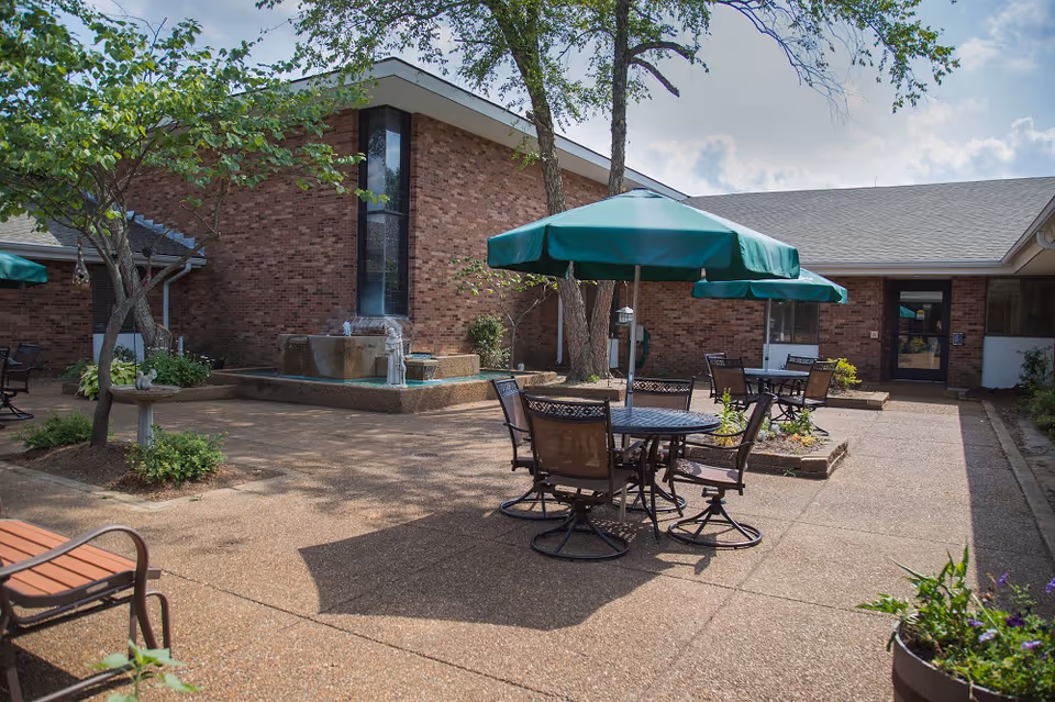 Outdoor courtyard area at St. Andrew's at Francis Place featuring round tables with green umbrellas, metal chairs, a water fountain, trees, and brick building walls under a partly cloudy sky.