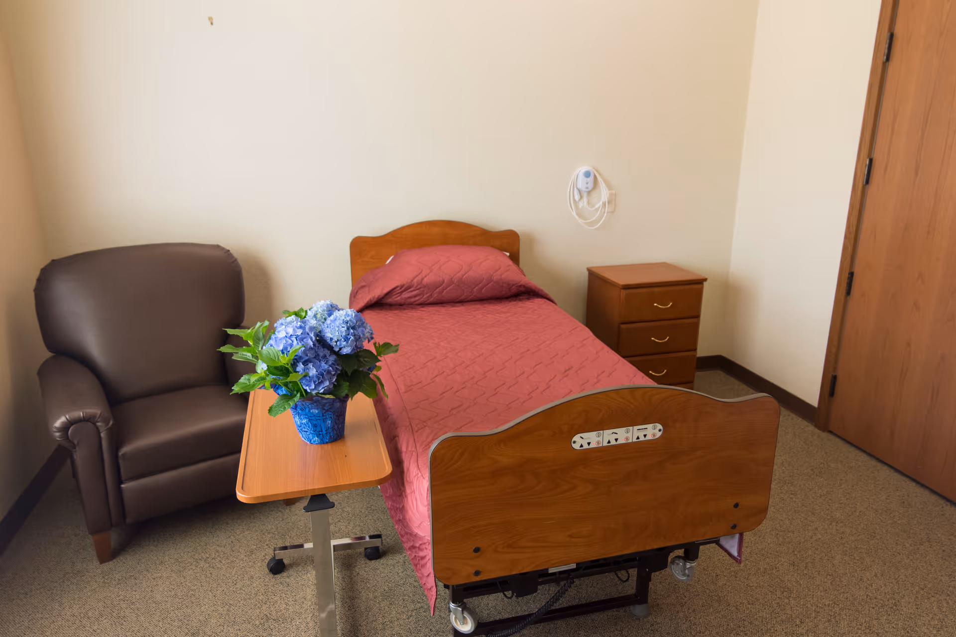 A small, simple bedroom in a senior living facility featuring a single hospital-style bed with a red quilted bedspread, a wooden nightstand with three drawers, a brown upholstered armchair, and a small overbed table with a blue vase holding blue hydrangea flowers. The walls are plain and light-colored, and there is a wooden door on the right side.