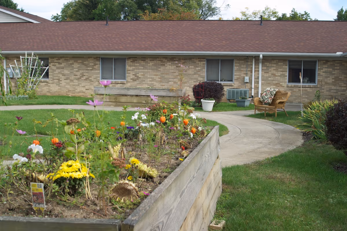 Outdoor garden area with raised flower beds containing various colorful flowers, a curved concrete pathway, green grass, and a wicker chair with a floral cushion near a brick building with several windows.