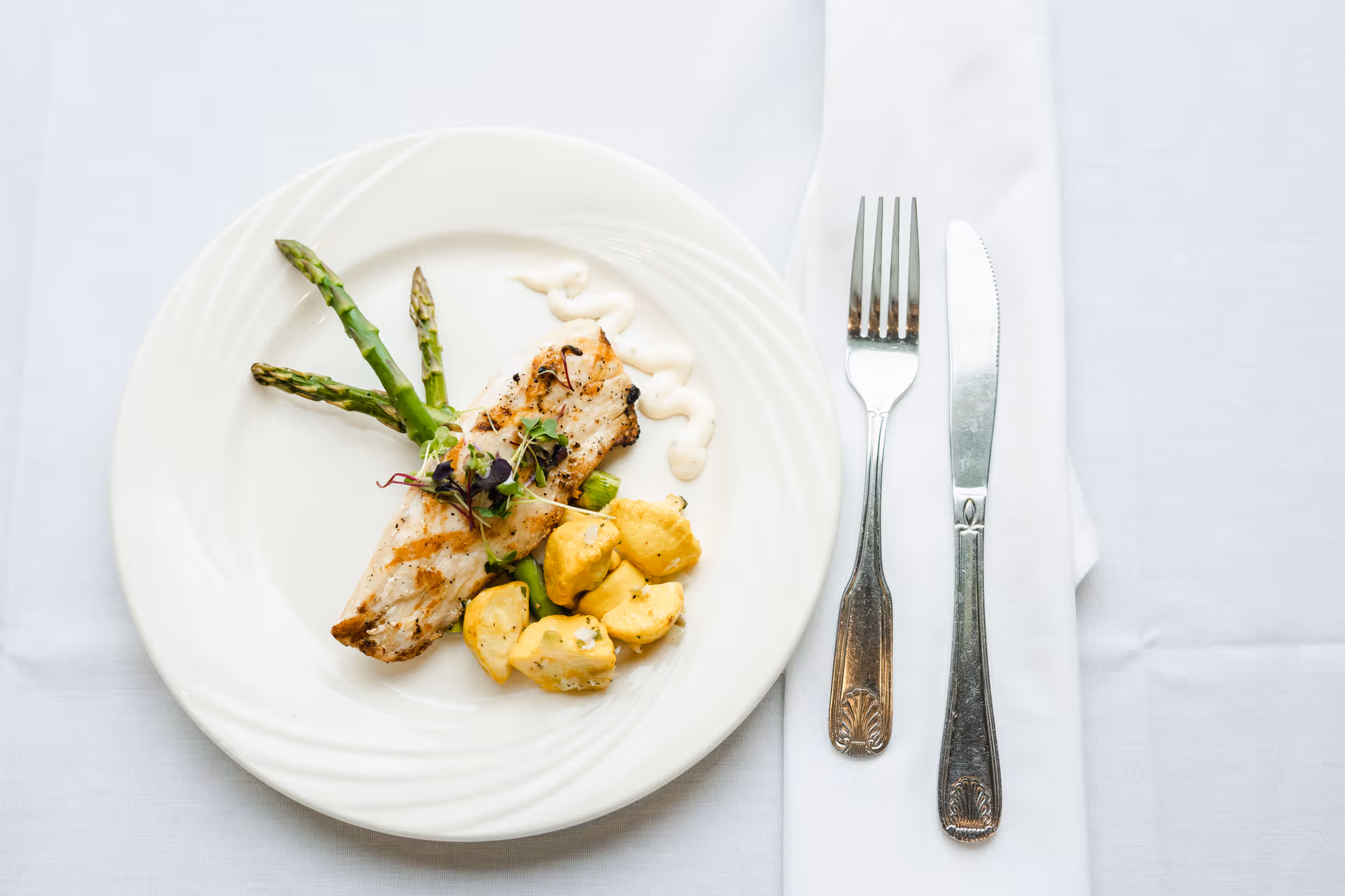 A white plate with grilled chicken breast, asparagus spears, and roasted potatoes garnished with microgreens and a drizzle of white sauce, placed on a white tablecloth next to a fork and knife on a folded white napkin.