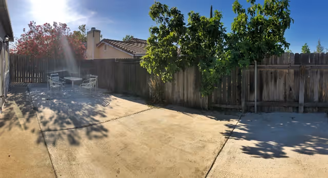 Outdoor patio area with concrete flooring, a wooden fence surrounding the space, a small table with four metal chairs, some trees and plants casting shadows, and a clear blue sky with sunlight shining.