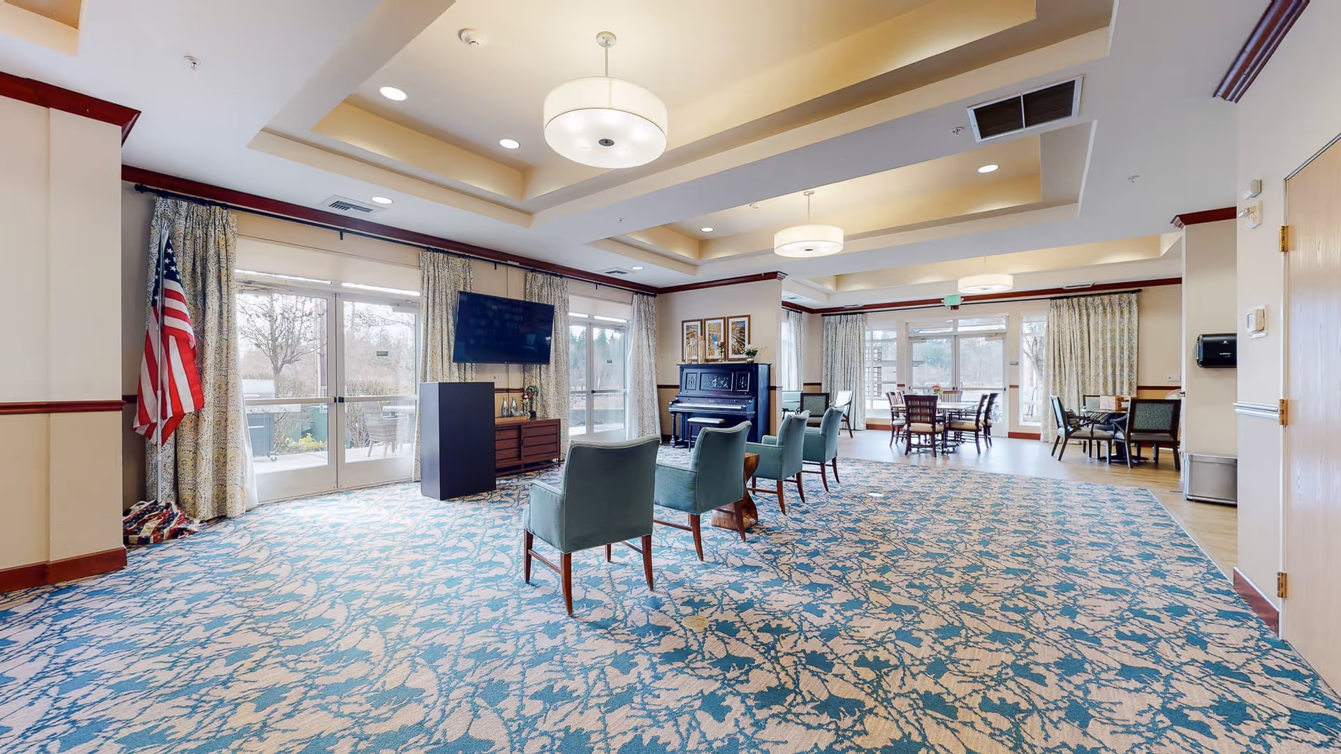 A spacious, well-lit common area in a senior living facility with patterned blue and beige carpet, several green upholstered chairs arranged in a semi-circle, a piano against the wall, a flat-screen TV mounted above a cabinet, and multiple tables and chairs near large windows and glass doors leading outside. The room features beige walls with wooden trim, ceiling lights, and an American flag in the corner.