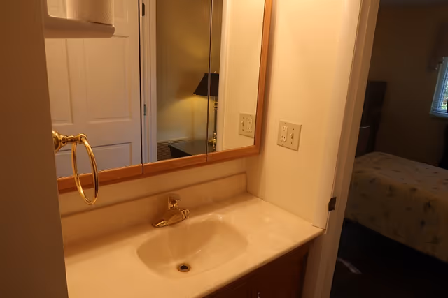 A bathroom sink with a beige countertop and a gold faucet. Above the sink is a large mirror with a wooden frame. To the left, there is a gold towel ring attached to the wall. In the reflection of the mirror, a lamp and part of a room with a bed and window are visible.