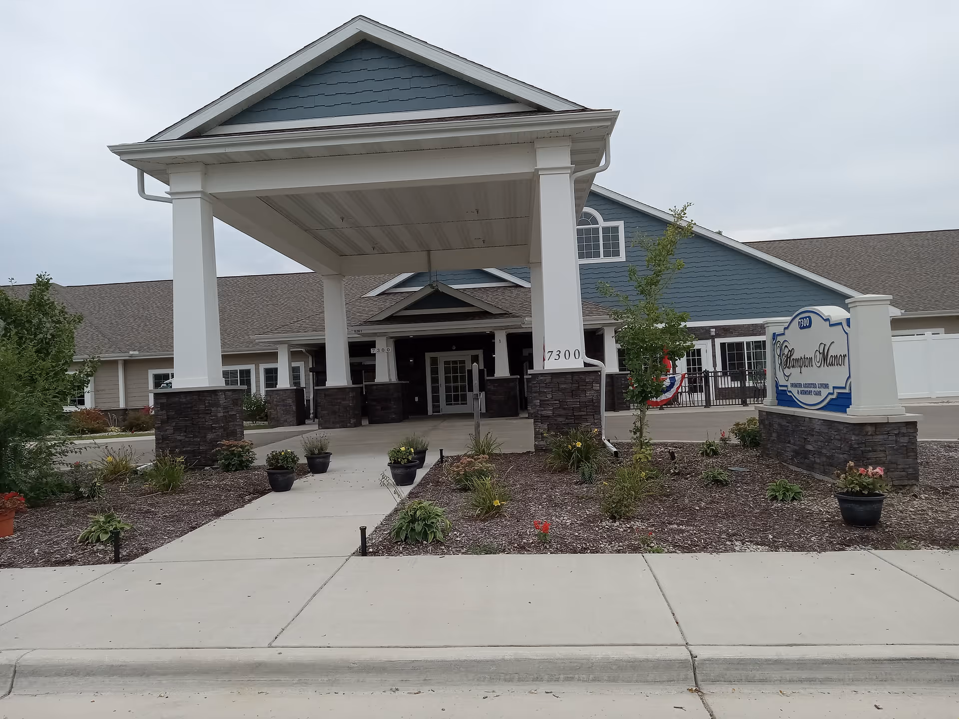 Front entrance of Hampton Manor assisted living building with a large covered porte-cochere, landscaped beds, and a facility sign near the sidewalk.