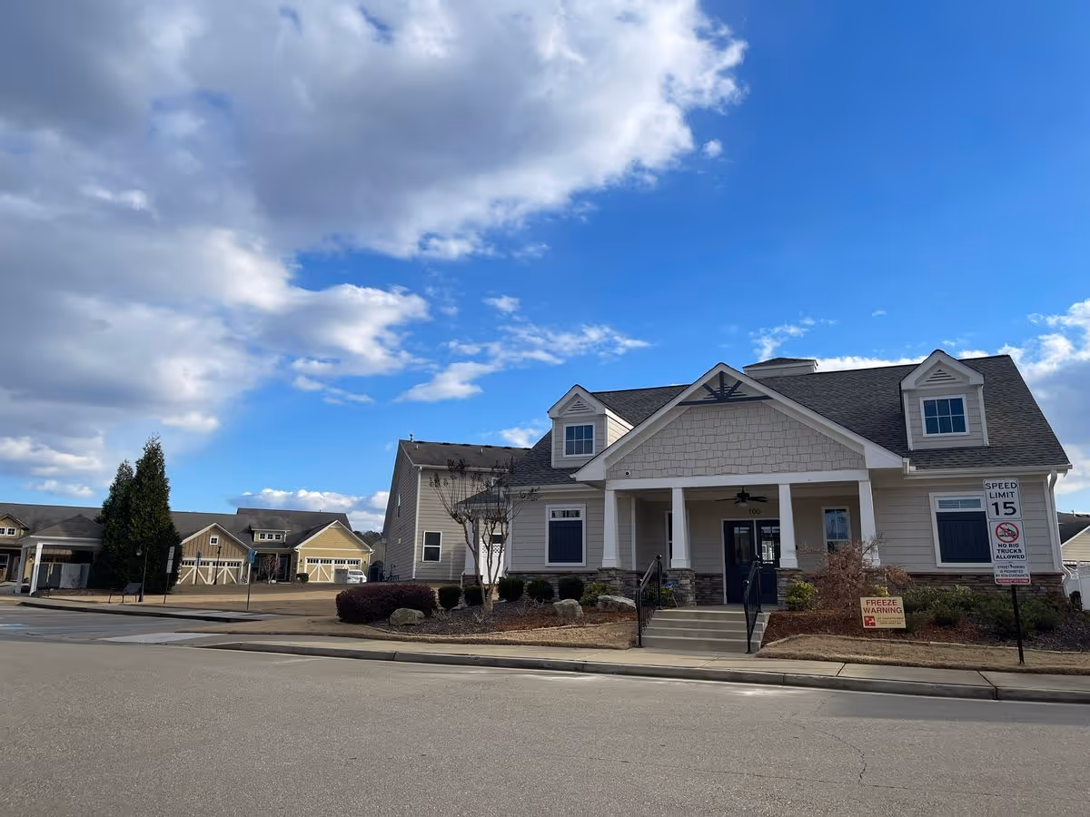 Front exterior of a residential community building with a pitched roof, covered porch entrance, and nearby garage-style units under a partly cloudy blue sky.