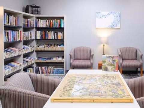 A cozy reading and puzzle area in a senior living facility featuring a large table with a partially completed jigsaw puzzle, surrounded by four striped upholstered chairs. Behind the table is a bookshelf filled with books and two additional striped chairs with a small side table and lamp between them. A framed artwork hangs on the wall above the lamp.