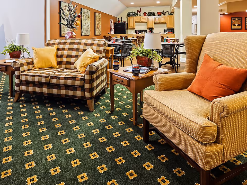 A cozy living room area in an assisted living facility featuring a checkered loveseat with two yellow pillows, a beige armchair with an orange pillow, a wooden side table with a lamp and plant, and a patterned green carpet. In the background, there is a dining area with tables and chairs and a kitchen with wooden cabinets and decorative items on top.