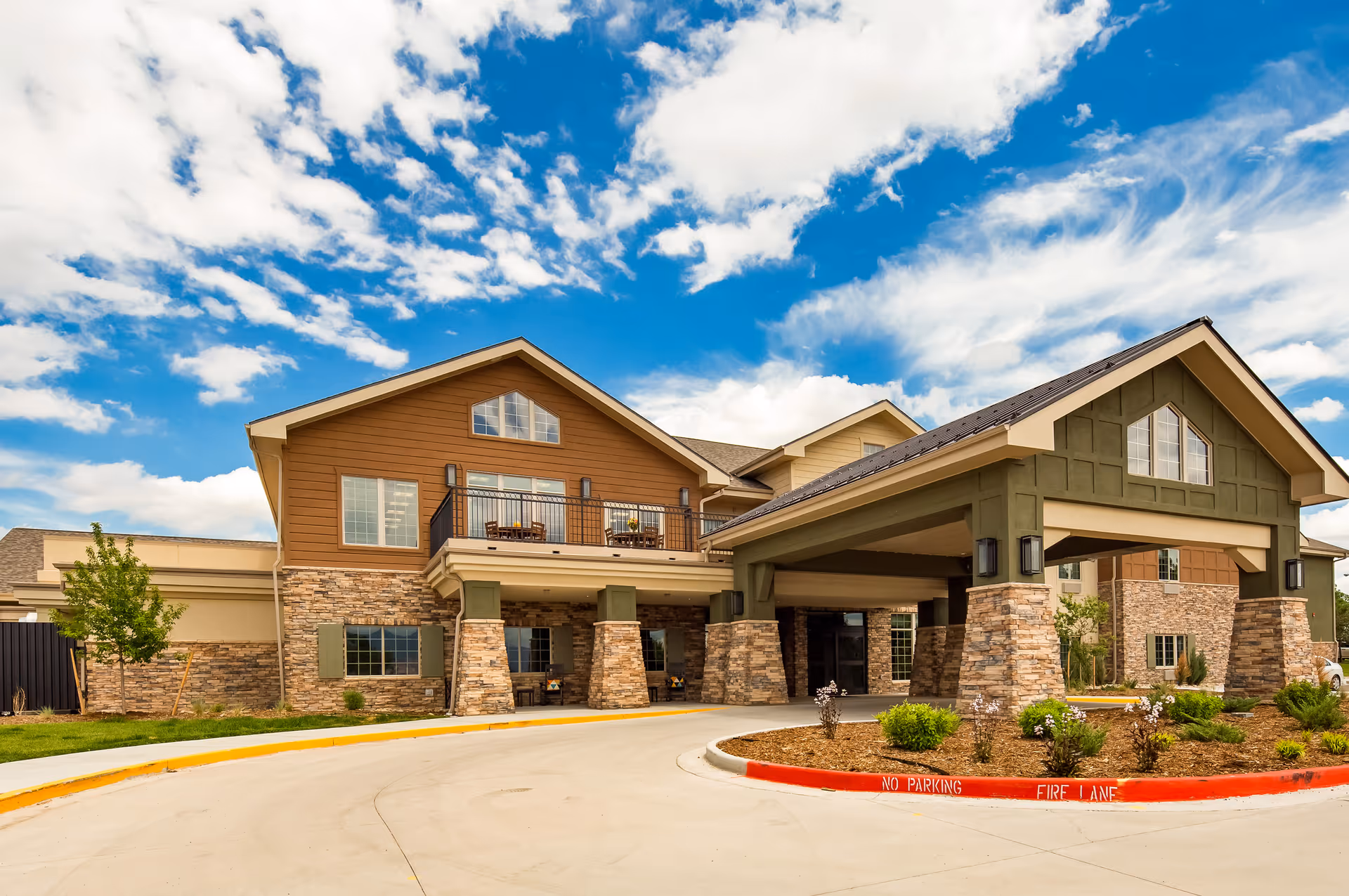 Exterior view of a senior living facility building with stone and wood siding, a covered entrance supported by stone pillars, and a balcony with outdoor seating under a partly cloudy blue sky.