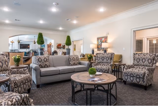 A well-lit senior living facility common area featuring a gray sofa with patterned cushions, four patterned armchairs, and a round wooden coffee table with a small green plant and magazines. The room has beige walls, carpeted flooring, decorative plants, table lamps, and artwork on the walls. In the background, there is a dining area with tables and chairs and a large mirror reflecting the space.