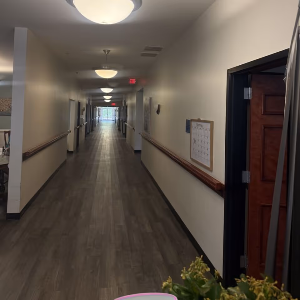 A long, well-lit hallway in a senior living facility with wood-look flooring, beige walls, handrails on both sides, ceiling lights, and an exit sign at the far end. There is a calendar on the right wall and a partially open wooden door on the right side near the foreground.