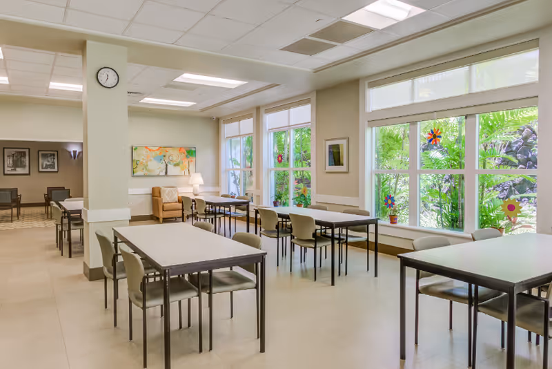 A bright and clean dining area with several tables and chairs arranged neatly. Large windows on one side let in natural light and show green foliage outside. The walls are decorated with framed artwork and colorful flower decals on the windows. A clock is mounted on a pillar in the room.