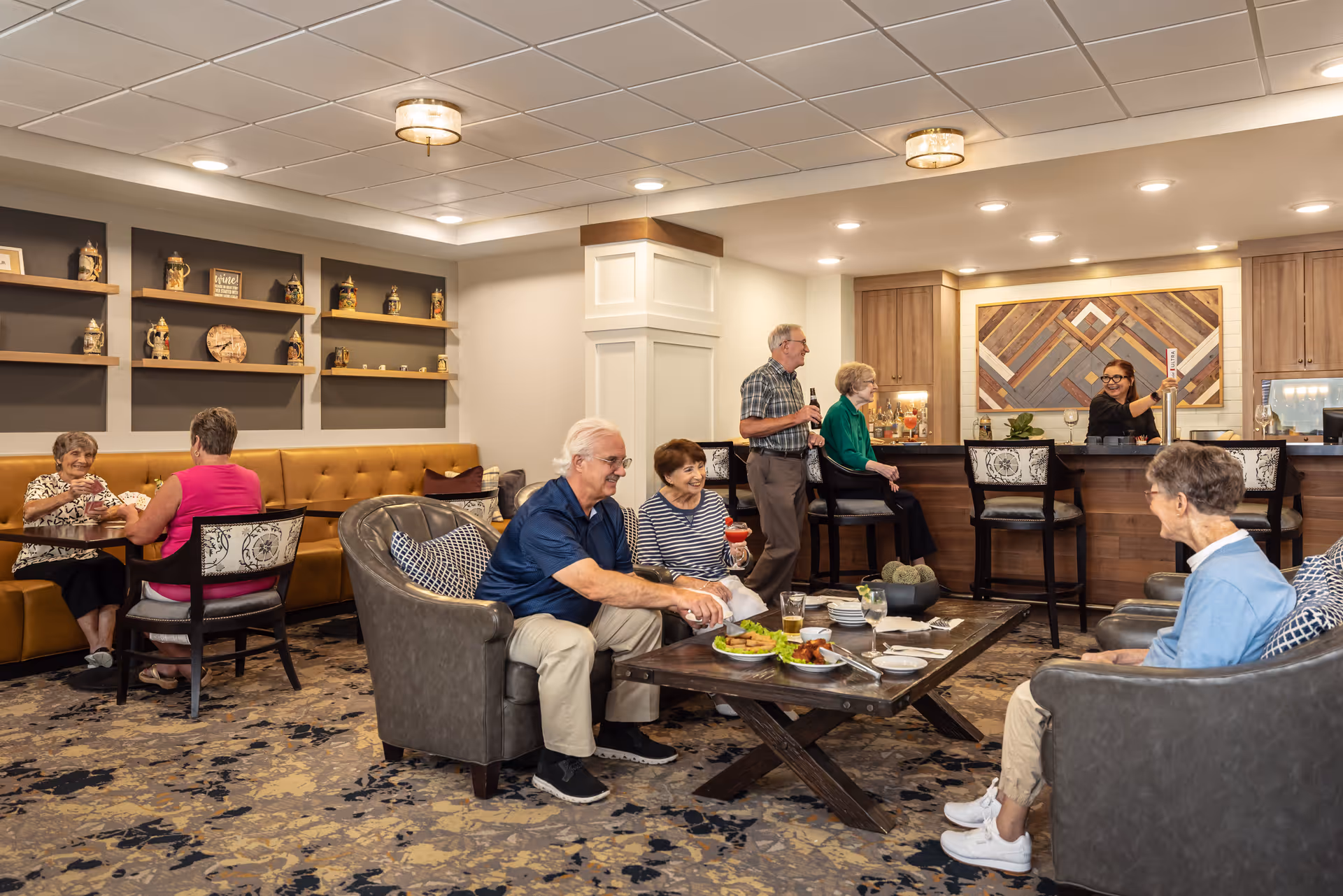 A cozy senior living facility lounge area with several elderly people socializing. Some are seated on comfortable chairs around a wooden coffee table with food and drinks, while others sit at a bar counter where a bartender is serving. The room features warm lighting, decorative shelves with pottery, and a patterned carpet.
