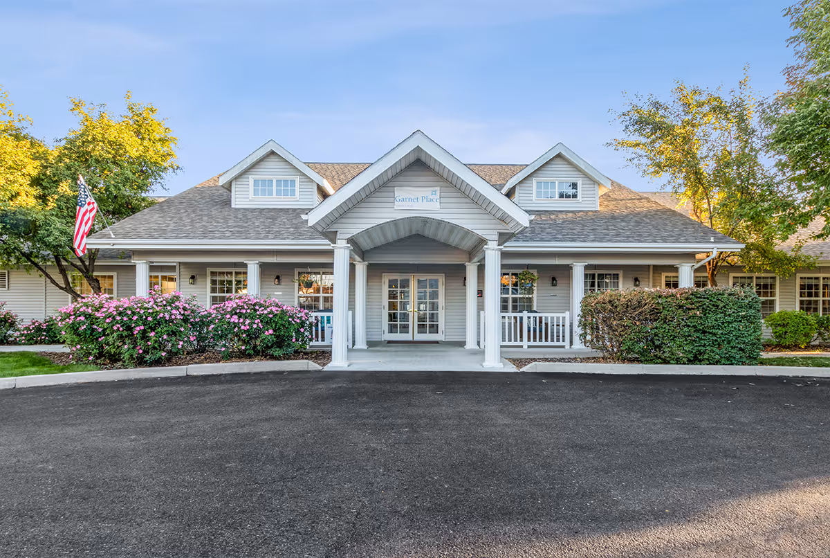 Front exterior view of a single-story senior living facility building with a covered entrance supported by white columns. The building has gray siding, multiple windows, and dormer windows on the roof. There are bushes with pink flowers and green trees around the entrance, and an American flag is displayed on the left side.