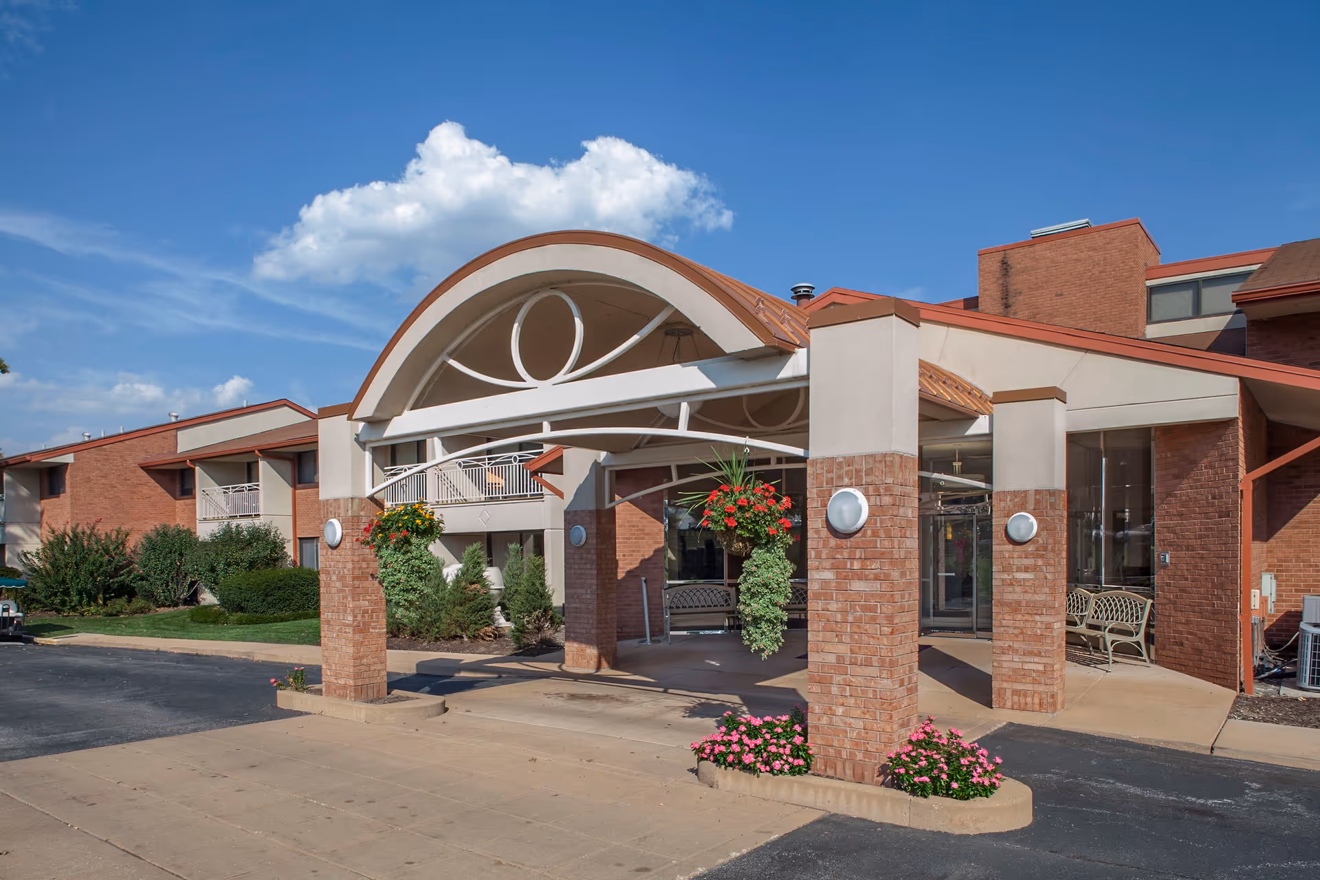 Exterior view of the entrance to a senior living facility named Garden Villas, featuring a covered driveway with brick pillars, hanging flower baskets, and a clear blue sky with some clouds.