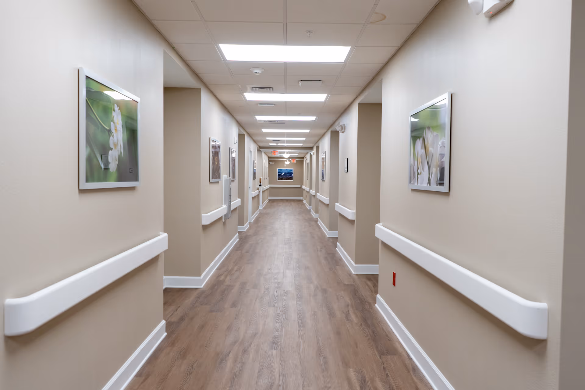 A long, well-lit hallway in a senior living facility with beige walls, wood flooring, white handrails on both sides, and framed floral artwork hanging on the walls.
