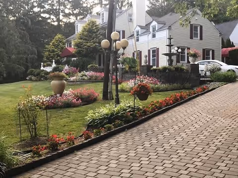 A paved driveway leading up to a white multi-story building with dark shutters, surrounded by a well-maintained garden featuring various flowers, shrubs, and decorative planters. There are lamp posts with multiple globes along the driveway, and a white car is parked near the building. Trees and greenery are visible in the background.