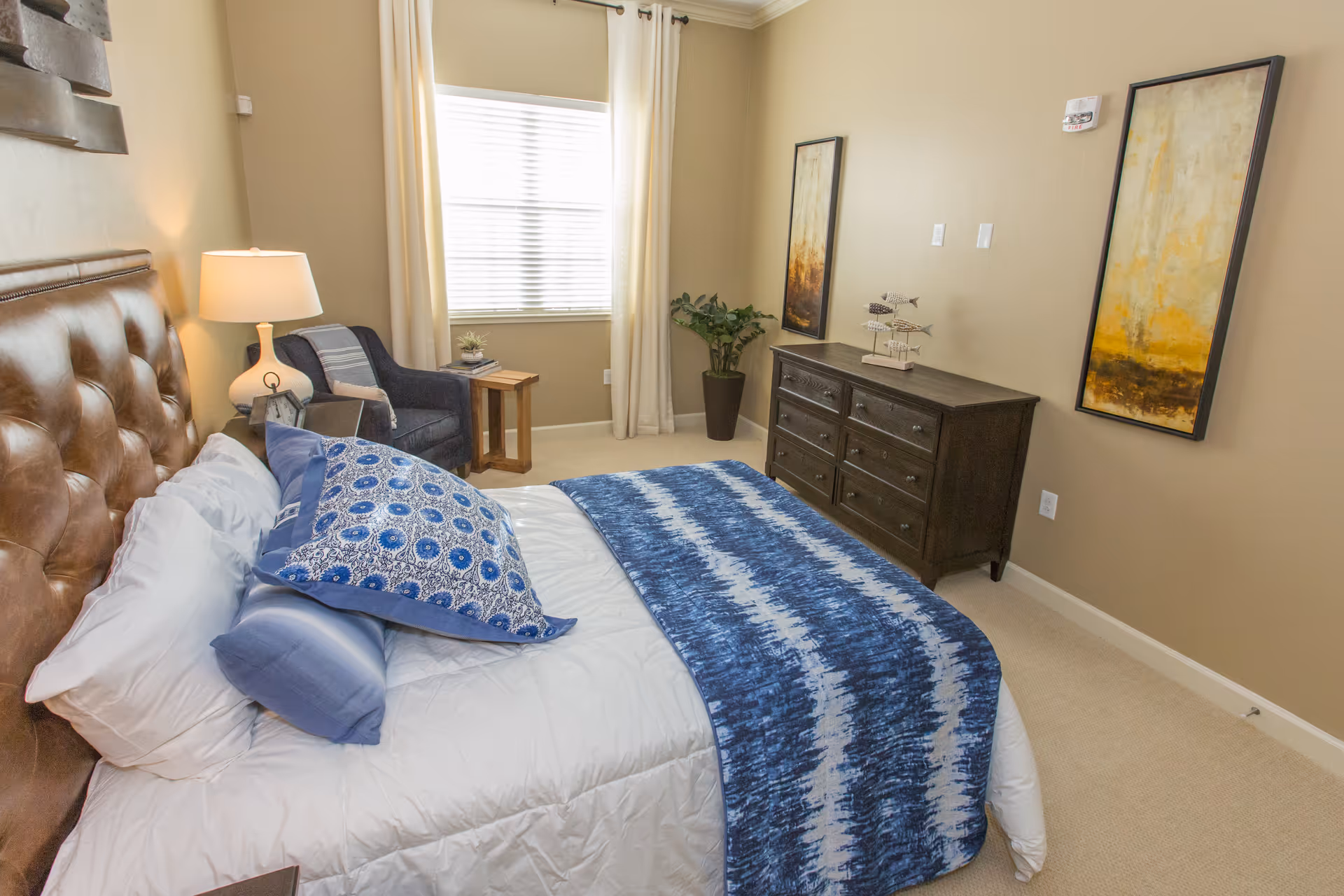 A cozy bedroom with a brown leather tufted headboard bed dressed in white bedding and blue patterned pillows and throw. There is a wooden nightstand with a white lamp and a small clock. A navy blue armchair with a striped throw blanket sits next to a wooden side table by a window with white curtains. Across from the bed is a dark wooden dresser with decorative fish sculptures and two abstract paintings on the beige walls. A potted plant is placed in the corner near the dresser.