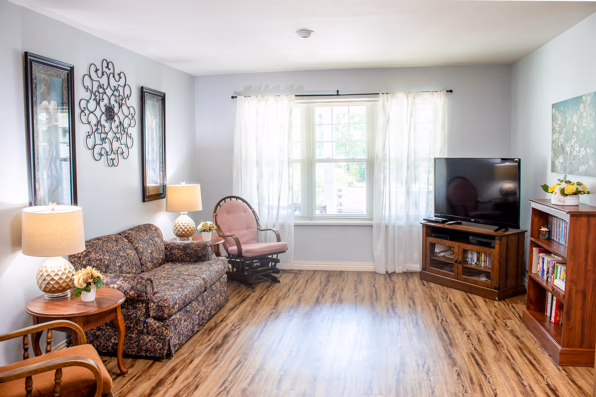 A bright living room with a floral-patterned sofa, a pink cushioned rocking chair, two side tables with lamps and small flower arrangements, a large window with sheer white curtains, a flat-screen TV on a wooden stand, and a wooden bookshelf filled with books. The room has light-colored walls and wood flooring.