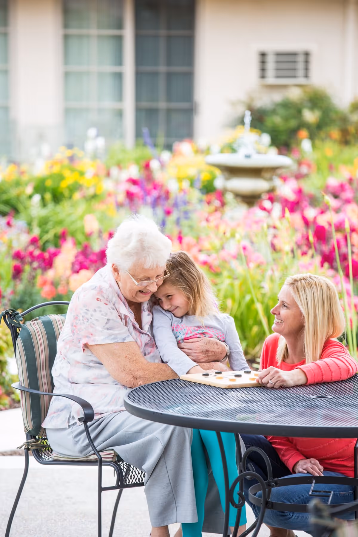 An elderly woman sitting at a round outdoor table with a young girl on her lap and a woman sitting next to them, all smiling and enjoying time together in a colorful garden with flowers and a fountain in the background.
