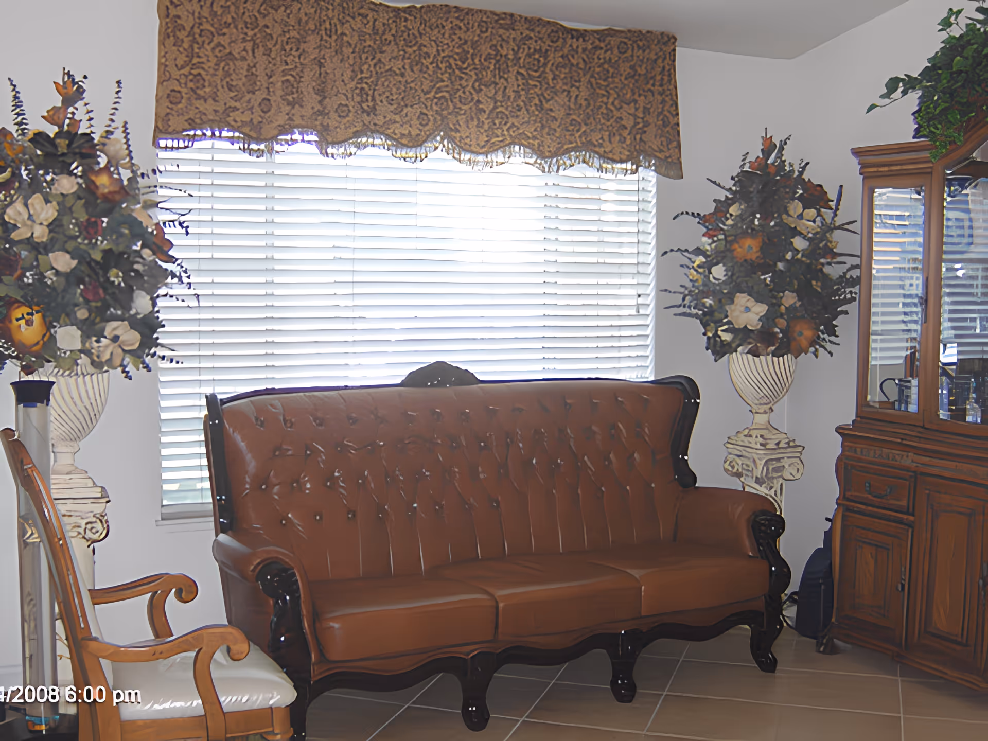 A living room with a brown tufted leather sofa, two large floral arrangements in white urns on either side of the sofa, a wooden chair with a white cushion, a wooden cabinet with glass doors on the right, and a window with white blinds and a patterned valance.