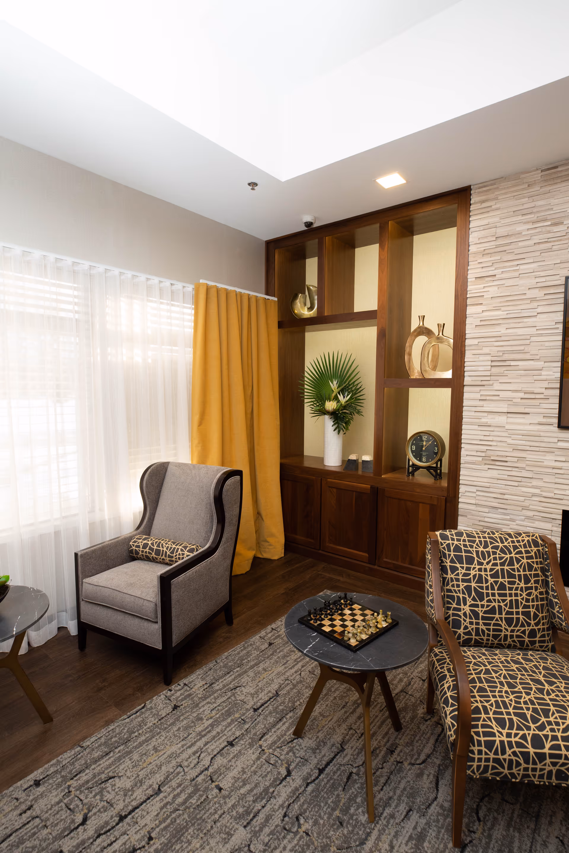 A cozy living room area with a gray armchair featuring a patterned cushion, a patterned upholstered chair, and a small round table with a chessboard on it. The room has a large window with sheer white curtains and mustard yellow drapes. There is a built-in wooden shelving unit with decorative items including a vase with green leaves and a clock. The floor is wooden with a textured area rug, and the wall has a light stone accent.