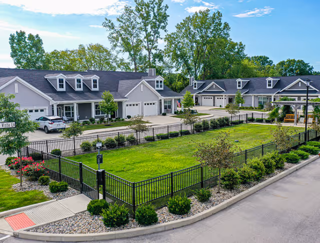 View of a residential senior living community with multiple single-story homes featuring garages, surrounded by a black metal fence enclosing a green lawn and landscaped bushes, under a blue sky with scattered clouds.