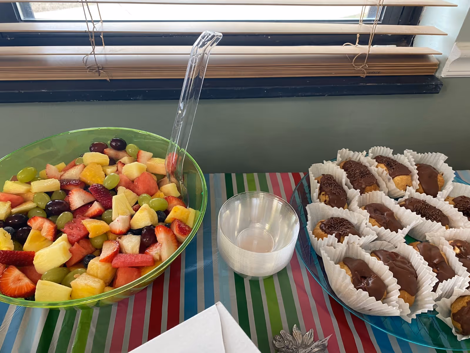 A green bowl filled with a colorful fruit salad containing strawberries, grapes, pineapple, and watermelon, next to a plate of chocolate-covered éclairs in white paper liners, with a stack of clear plastic bowls in between. The items are placed on a table covered with a striped tablecloth in blue, green, and pink colors, beneath a window with partially open blinds.