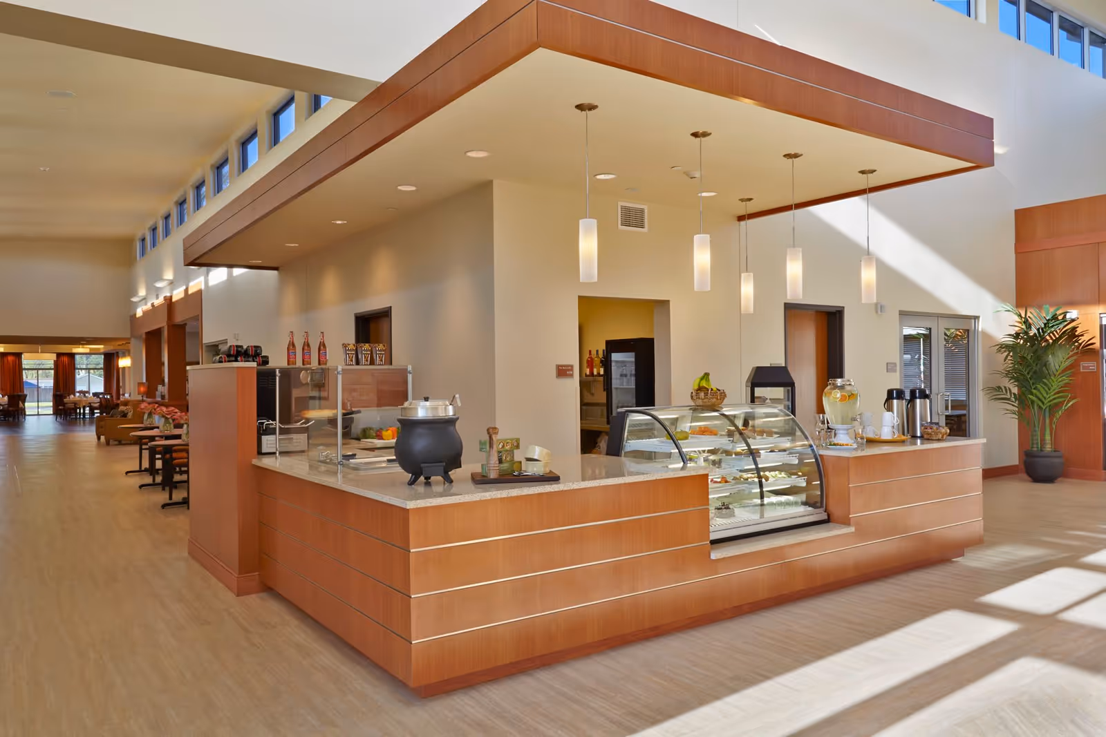 A bright and spacious interior area of Arlington Place Health Campus featuring a wooden counter with a glass display case containing desserts and fruits. There are hanging pendant lights above the counter, a beverage dispenser with cups, and a basket of bananas. The area extends into a hallway with seating and tables visible in the background, with large windows allowing natural light to fill the space.