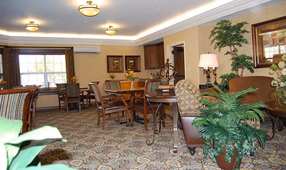 Well-lit common dining area with wooden tables and chairs, upholstered armchairs, decorative plants and framed artwork.
