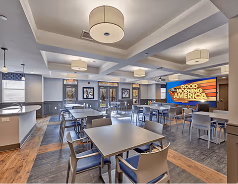 A bright and spacious dining room with multiple tables and chairs arranged neatly. The room features modern ceiling lights, large windows allowing natural light, and a large screen on the wall displaying the Good Morning America logo. The flooring is a mix of wood and tile, and the walls are painted in neutral tones with decorative trim.
