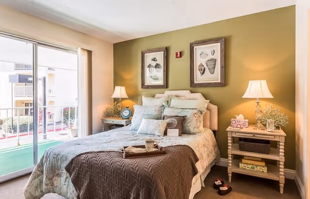 A cozy bedroom with a neatly made bed featuring multiple pillows and a brown throw blanket. Two bedside tables with lamps, decorative items, and framed artwork hang on a green accent wall behind the bed. A sliding glass door opens to a balcony with a view of the outside.